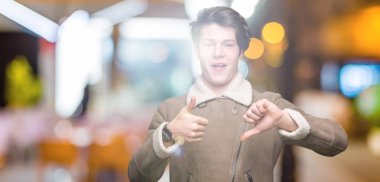 İzole arka plan üzerinde kış ceket giyen genç yakışıklı adam başparmak yukarı ve aşağı, anlaşmazlık ve anlaşma ifade yapıyor. Çılgın çatışma