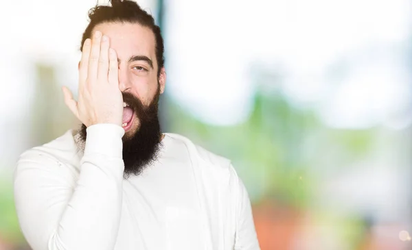 Young man with long hair and beard wearing sporty sweatshirt covering one eye with hand with confident smile on face and surprise emotion.