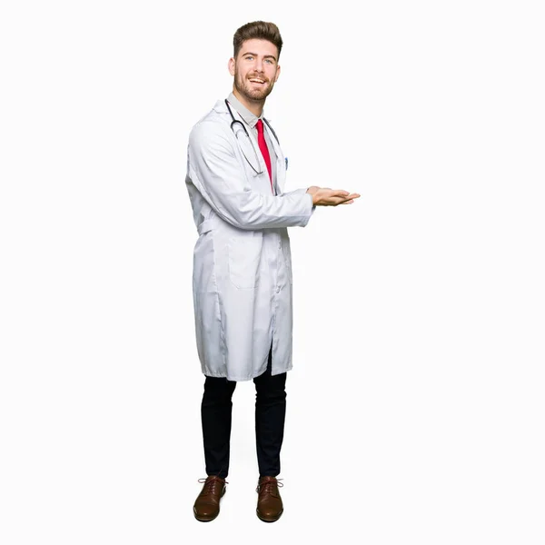 年轻英俊的医生穿着医疗外套指着身边的手和张开的手掌 呈现广告微笑的快乐和自信 — 图库照片