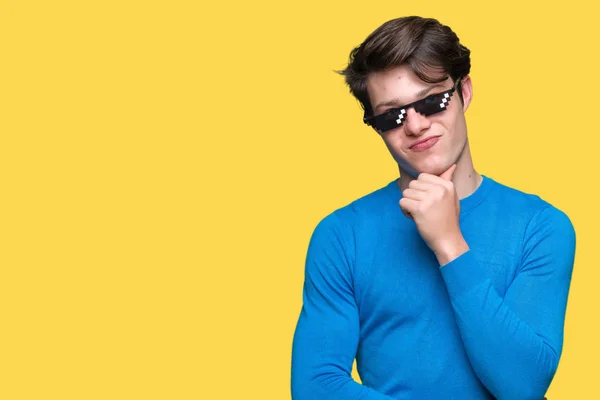 Young man wearing funny thug life glasses over isolated background with hand on chin thinking about question, pensive expression. Smiling with thoughtful face. Doubt concept.