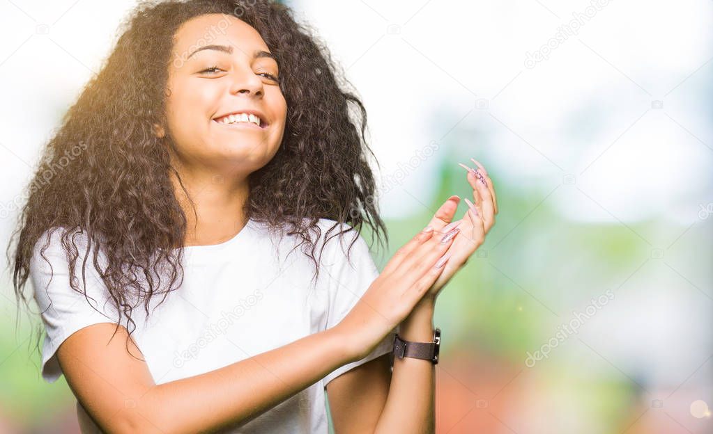 Young beautiful girl with curly hair wearing casual white t-shirt Clapping and applauding happy and joyful, smiling proud hands together