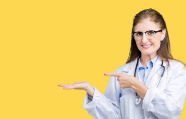 Orta yaş olgun doktor kadın hayrete ve el ile sunulması ve parmak ile işaret ederken kameraya gülümseyen izole arka plan üzerinde tıbbi bir palto giyiyor.