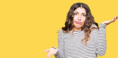 Çizgili kazak clueless ve şaşkın bir ifadeyle kollar ve eller kaldırdı giyen genç güzel kadın. Şüphe kavramı.