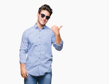 Yakışıklı delikanlı arıyor ve başparmak yanında yukarıyı mutlu yüz ile gülümseyen izole arka plan üzerinde güneş gözlüğü takıyor.