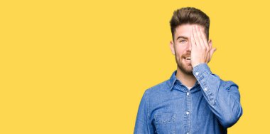 Genç yakışıklı sarışın adam el yüz ve sürpriz duygu kendinden emin gülümseme ile tek gözüyle kapsayan geçici kot gömlek giymiş.