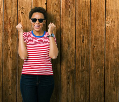 Güzel genç Afro-Amerikan kadın izole arka plan kutlama üzerinde güneş gözlüğü takıyor şaşırttı ve silah ile başarı için yükseltilmiş ve açık gözler hayrete düşürdü. Kazanan kavramı.