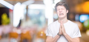 Yalvarıyor ve umut ifade yüzünde çok duygusal ve endişe ile birlikte elleriyle dua izole arka plan üzerinde rahat beyaz t-shirt giyen genç yakışıklı adam. Bağışlanmak. Din kavramı.