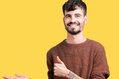 İzole arka plan gösteren palm el üzerinde kış kazak giyen ve yukarıya, mutlu ve neşeli gülümseyen baş parmakları Tamam jest yapıyor yakışıklı delikanlı