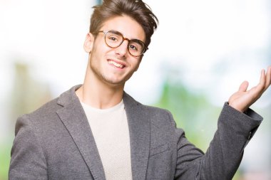 Neşeli sunulması ve kameraya bakarak avucunun ile işaret gülümseyen izole arka plan üzerinde gözlük takan genç iş adamı.