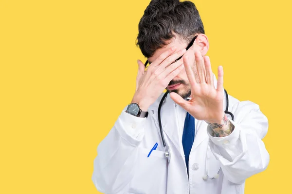 Young doctor man wearing hospital coat over isolated background covering eyes with hands and doing stop gesture with sad and fear expression. Embarrassed and negative concept.