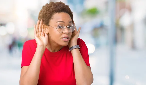 Beautiful young african american woman wearing glasses over isolated background Trying to hear both hands on ear gesture, curious for gossip. Hearing problem, deaf