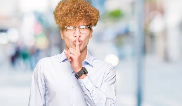 Young handsome business man with afro wearing glasses asking to be quiet with finger on lips. Silence and secret concept.