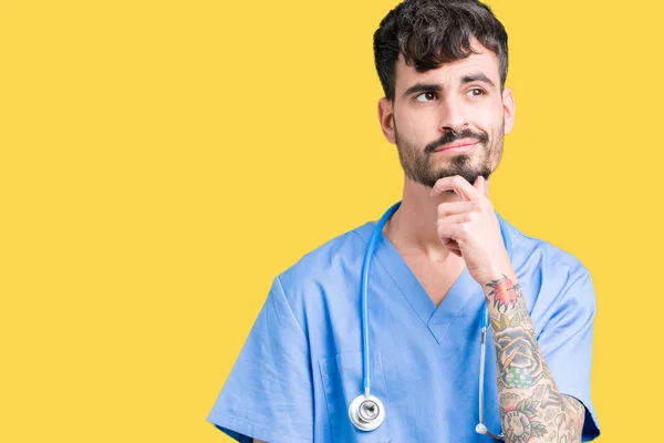 Young handsome nurse man wearing surgeon uniform over isolated background with hand on chin thinking about question, pensive expression. Smiling with thoughtful face. Doubt concept.