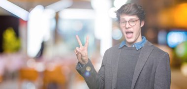 Mutlu yüz zafer işareti yaparken kameraya göz kırpıyor ile gülümseyen izole arka plan üzerinde gözlük takan genç yakışıklı iş adamı. İki numaralı.