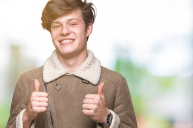 Yakışıklı delikanlı izole arka plan başarı işareti el ile pozitif jest yapıyor üzerinde kış palto giyiyor başparmak gülümseyen ve mutlu. Neşeli bir ifadeyle, kazanan jest kameraya bakıyor.