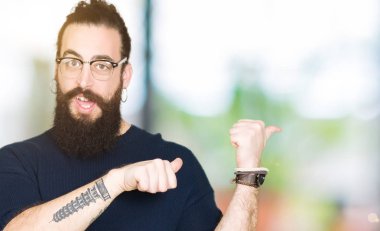 Uzun saç ve sakal giyen genç hipster adamla emin gülümseyen işaretleme için arkasından el ve başparmak yukarıya, gözlük