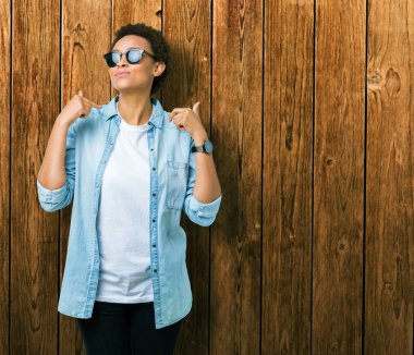 Güzel genç Afro-Amerikan kadın yüzünde gülümseme ile kendinden emin görünüyorsun, kendini gururlu ve mutlu parmakları ile işaret izole arka plan üzerinde güneş gözlüğü takıyor.