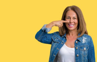 Casual denim ceket izole üzerinde giyen güzel orta yaş kadın arka plan işaretçi el parmak yüz ve burun, neşeli gülümseyen