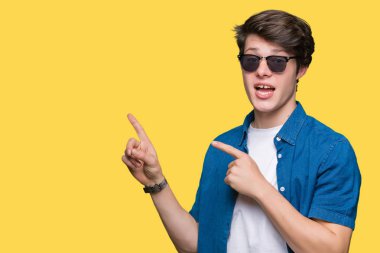 Yakışıklı delikanlı gülümseyen ve iki elleri ve parmakları ile tarafına işaret kamera bakarak izole arka plan üzerinde güneş gözlüğü takıyor.