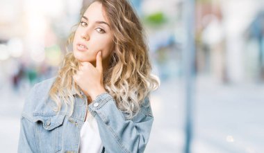 Emin çapraz kollar ve çeneme kaldırdı el ile gülümseme ile kameraya bakarak izole arka plan üzerinde kot ceket giyen güzel genç sarışın kadın. Pozitif düşünce.