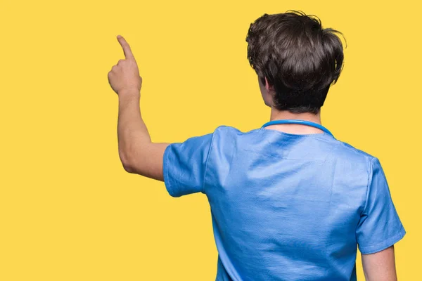 年轻医生穿着医疗服在孤立的背景姿势向后指着背后用手指的手 — 图库照片