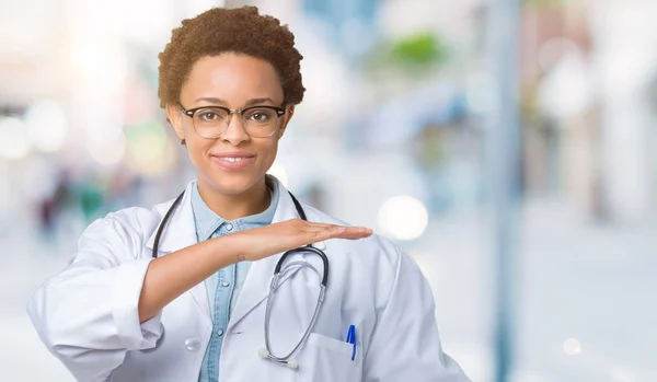 Young african american doctor woman wearing medical coat over isolated background gesturing with hands showing big and large size sign, measure symbol. Smiling looking at the camera. Measuring concept.