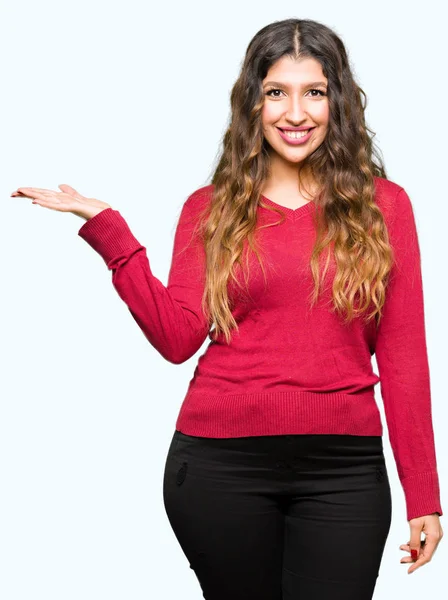 陽気な提示とカメラを見て手の手のひらで指している笑みを浮かべて赤いセーターを着ている若い美しい女性 — ストック写真
