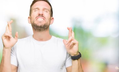 Rahat beyaz t-shirt giyen yakışıklı adam umut ve gözler kapalı parmaklar geçiş gülümseyen. Şans ve batıl fikir.