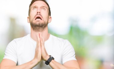 Yakışıklı adam yalvarıyor ve umut ifade yüzünde çok duygusal ve endişe ile birlikte elleriyle dua casual beyaz t-shirt giyiyor. Bağışlanmak. Din kavramı.