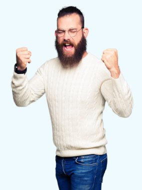 Gülümseyen ve başarı için çığlık gözlük ve kış kazak çok mutlu ve heyecanlı kaldırdı, silah ile kazanan jest yapıyor giyen genç hipster adam. Kutlama kavramı.