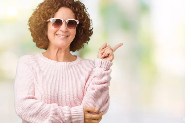 美しい中間エイガー年配の女性の顔に大きな笑みを浮かべて孤立の背景にピンクのセーター サングラスを着てカメラを見て側の手と指で指す — ストック写真
