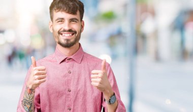 Yakışıklı delikanlı izole arka plan başarı işareti el ile pozitif jest yapıyor üzerinde pembe gömlek giymiş başparmak gülümseyen ve mutlu. Neşeli bir ifadeyle, kazanan jest kameraya bakıyor.