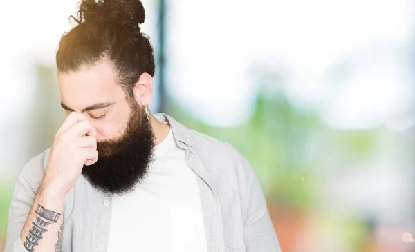 Young man with long hair, beard and earrings tired rubbing nose and eyes feeling fatigue and headache. Stress and frustration concept.