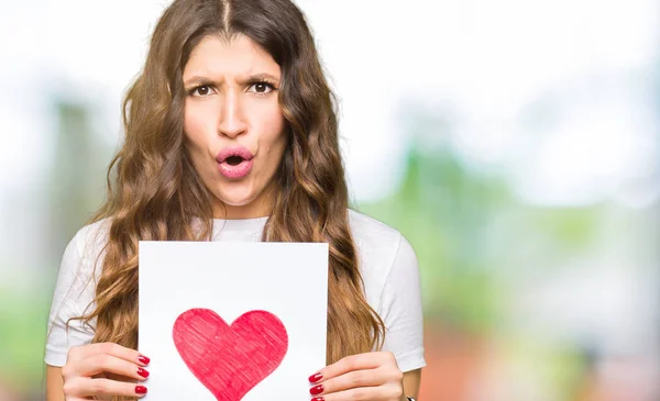 Young adult woman holding card with red heart scared in shock with a surprise face, afraid and excited with fear expression