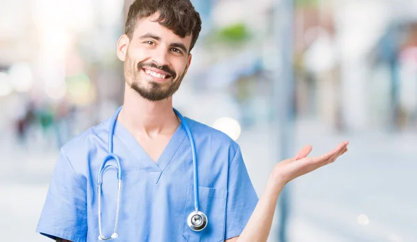 Young handsome nurse man wearing surgeon uniform over isolated background smiling cheerful presenting and pointing with palm of hand looking at the camera.
