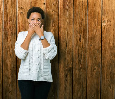 İzole arka plan üzerinde kazak giyen güzel genç Afrikalı Amerikalı kadın hata elleri ile ağız kapsayan şok. Gizli konsept.