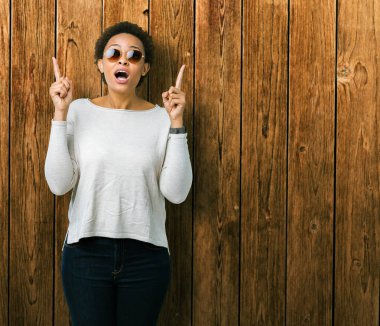 Güzel genç Afro-Amerikan kadın hayrete ve yukarıya bakıyor ve parmakları ile işaret ve silah kaldırdı sürpriz izole arka plan üzerinde güneş gözlüğü takıyor.