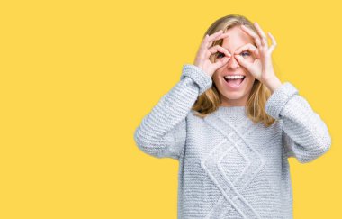 Dilini, parmaklarının arasından baktığını yapışmasını dürbün gibi ok jest yapıyor izole arka plan üzerinde kış kazak giyen güzel genç kadın. Deli ifade.