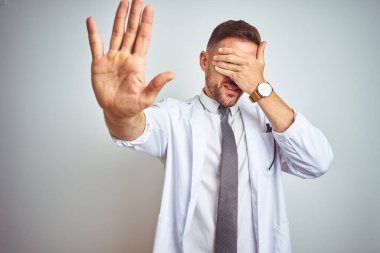 Genç yakışıklı Doktor adam el ile gözler kapsayan izole arka plan üzerinde beyaz profressional ceket giyiyor ve üzücü ve korku ifadesi ile stop jest yapıyor. Utanç verici ve negatif kavram.