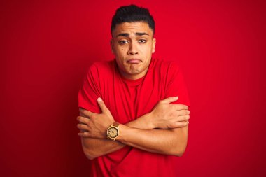 Yüz üzerinde üzücü ve şok ifade ile kış soğuk için sallayarak ve donma izole kırmızı arka plan üzerinde ayakta t-shirt giyen genç Brezilyalı adam