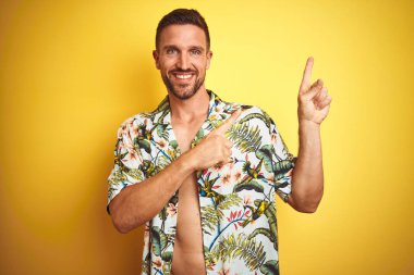 Sarı izole arka plan üzerinde yaz Hawaii çiçekler gömlek giyen yakışıklı adam gülümseyen ve yan iki el ve parmak ile işaret kamera bakıyor.