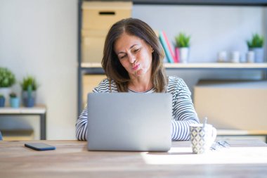 Orta yaş kıdemli kadın eğlenceli yüz ile bilgisayar laptop kabarık yanakları kullanarak evde çalışma masasında oturuyor. Ağız hava ile şişirilmiş, çılgın ifade.
