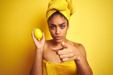 Afro amerikan kadın kamera ve size parmak ile işaret izole sarı arka plan üzerinde limon tutan duş tan sonra havlu giyen, el işareti, önden olumlu ve kendinden emin jest