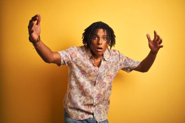 Tatil dreadlocks ile Afro adam izole sarı arka plan üzerinde çiçek gömlek giyiyor korkuyor ve sürpriz ifade, korku ve heyecanlı yüz ile şok.