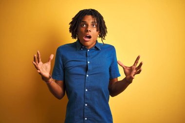 Çılgın ve deli çığlık ve agresif ifade ve silah kaldırdı ile bağırarak izole sarı arka plan üzerinde ayakta rahat denim gömlek giyen dreadlocks ile Afro adam. Hayal kırıklığı kavramı.