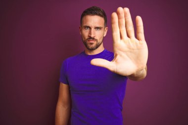 Lila izole arka plan üzerinde rahat mor t-shirt giyen genç adam avuç içi ile şarkı durdurmak yapıyor. Yüzünde olumsuz ve ciddi bir jest olan uyarı ifadesi.