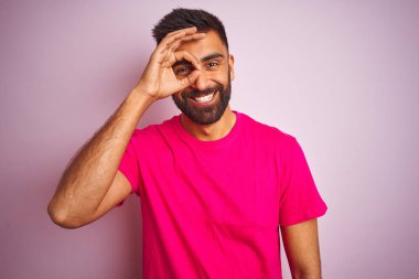 İzole pembe arka plan üzerinde duran t-shirt giyen genç hintli adam el gülümseyerek ok jest yapıyor, mutlu yüz ile parmaklardan bakan göz.