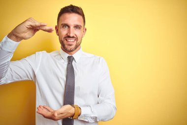 Genç yakışıklı iş adamı sarı izole arka plan üzerinde zarif beyaz gömlek giyen elleri büyük ve büyük boy işareti gösteren gesturing, ölçü sembolü. Kameraya bakarak gülümsüyor. Ölçme kavramı.