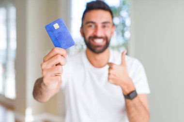 Yakışıklı İspanyol adam ok işareti yapıyor büyük bir gülümseme ile kredi kartı mutlu tutan, parmakları ile başparmak, mükemmel işareti