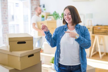 Genç çift yuvarlak karton kutular yeni bir eve hareket, artı boyutu kadın evde gülümseyerek ayakta ve yan iki el ve parmakları ile işaret kamera bakarak.